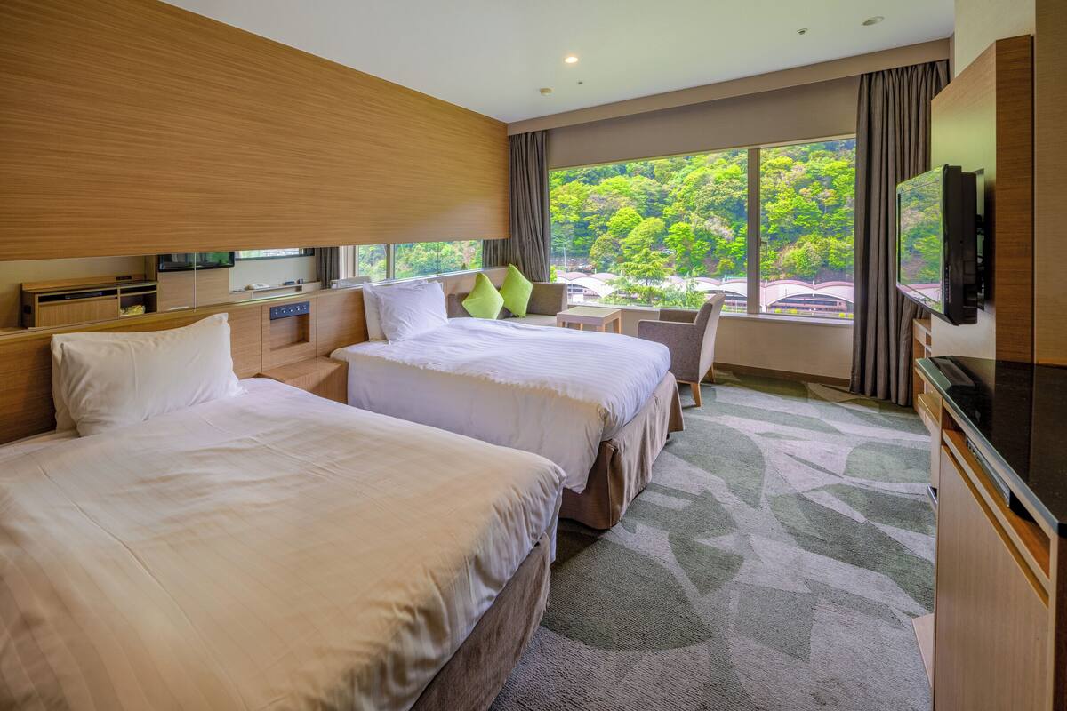 Quarto de hotel com duas camas, uma televisão, uma escrivaninha e vista para árvores através de amplas janelas.