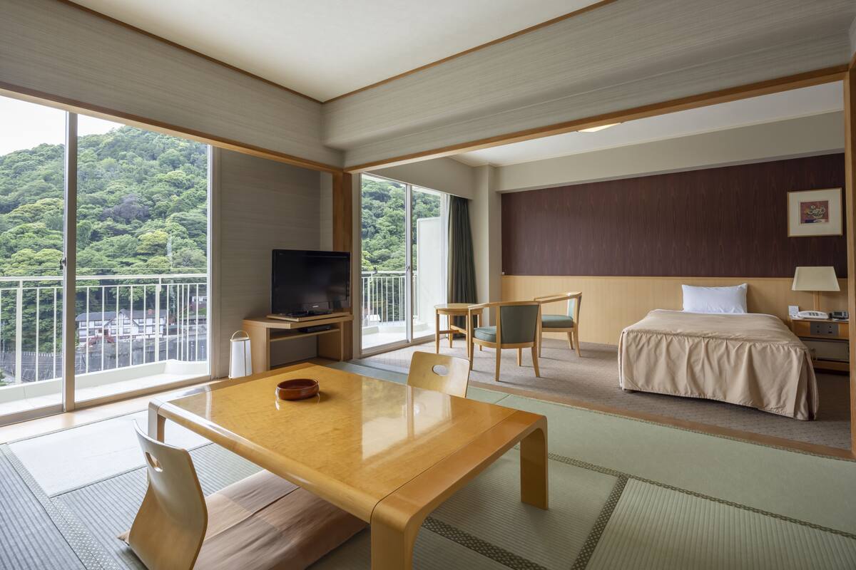 Quarto de hotel com uma mesa de jantar de madeira, uma cama com colcha bege, uma televisão e uma grande janela com vista para uma área arborizada.