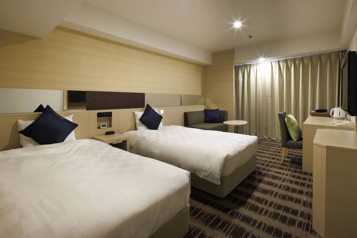 Quarto de hotel com duas camas, uma escrivaninha, uma cadeira, uma mesinha, uma televisão e uma janela com cortinas.