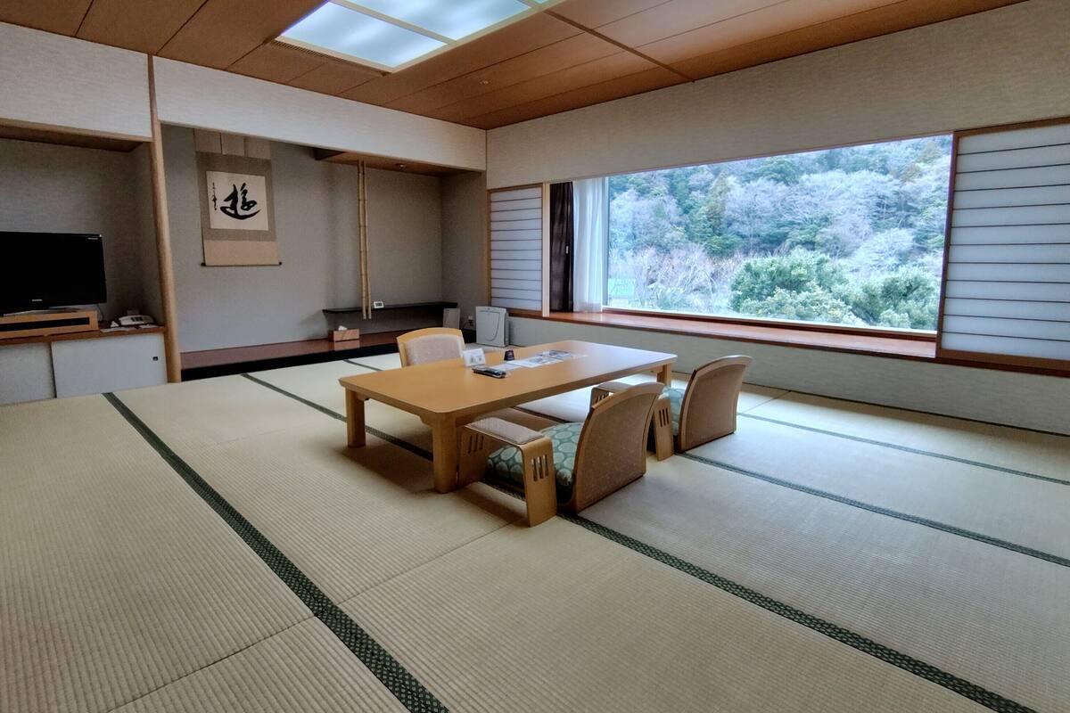 Um quarto com esteiras de tatame, uma mesa de madeira e cadeiras, uma televisão e uma grande janela com vista para árvores.
