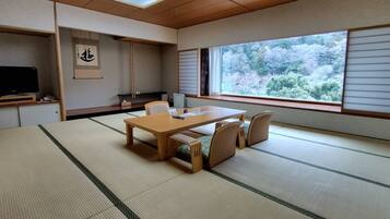 Een kamer met tatami matten, een houten tafel en stoelen, een televisie en een groot raam met uitzicht op bomen.