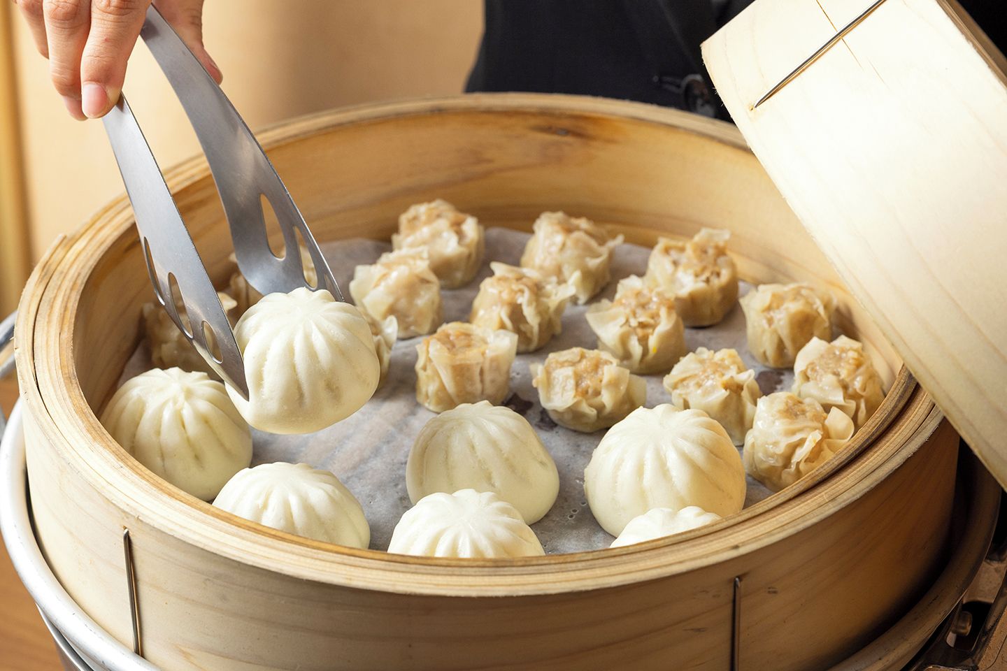 A person using tongs to handle dumplings in a bamboo steamer.