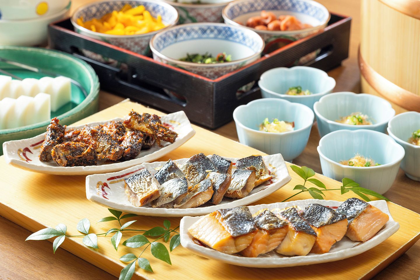 Diversos platos, entre ellos pescado a la parrilla y otras comidas, dispuestas sobre una mesa de madera con cuencos y platos.