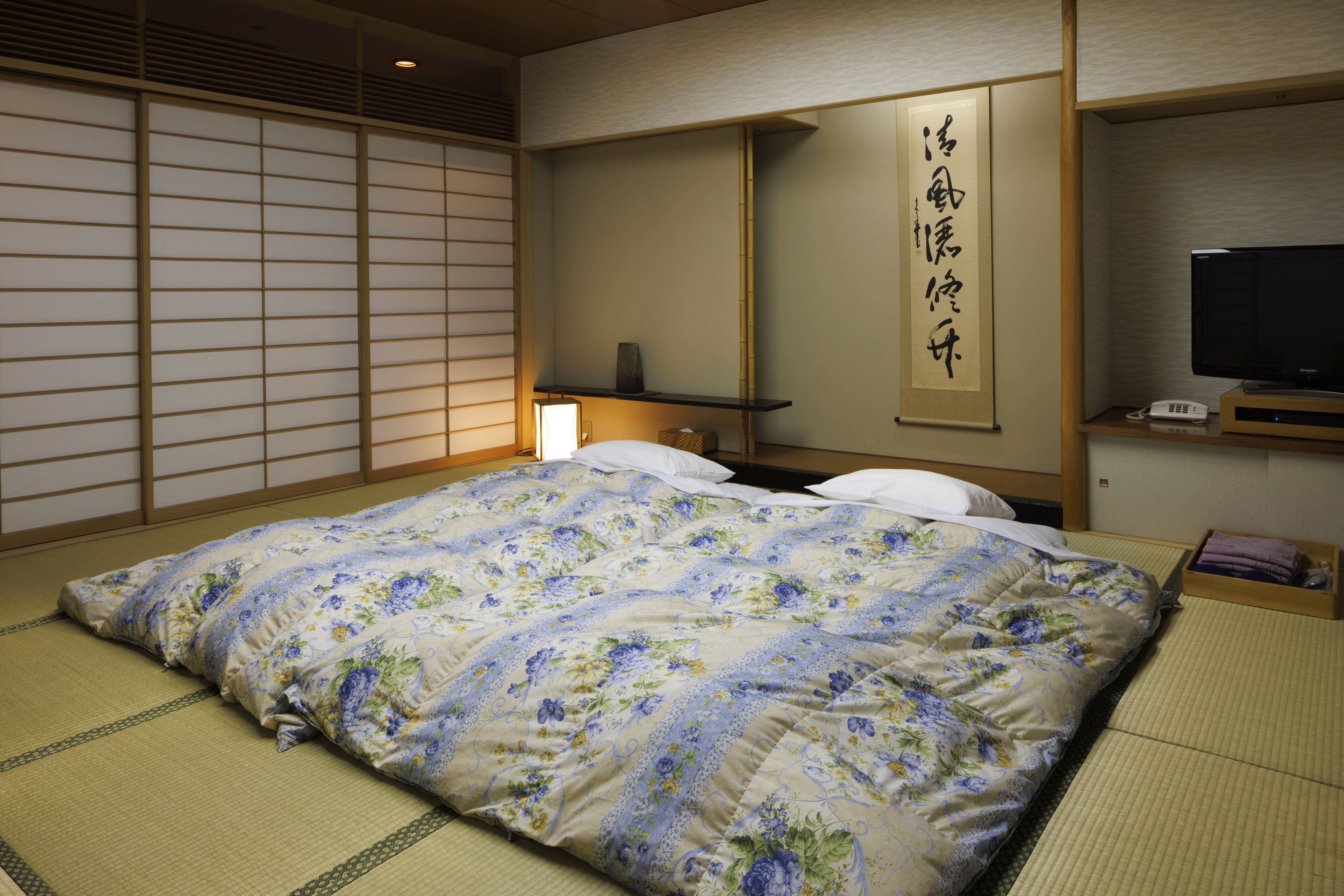 Ein traditioneller japanischer Raum mit Tatami-Boden, einem Bett mit floralem Überwurf, einem Fernseher und einem Regal mit einer Lampe.