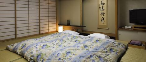 Una habitación japonesa tradicional con suelo de tatami, una cama con edredón floral, un televisor y una estantería con una lámpara.