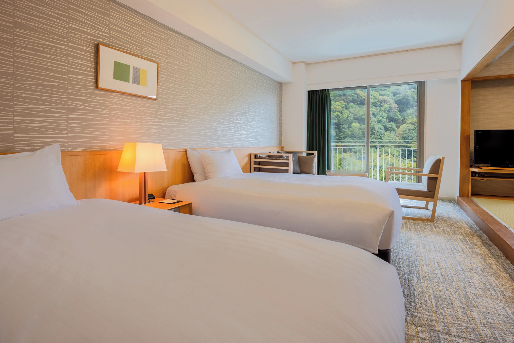 Habitación de hotel con dos camas, un escritorio, una silla, un televisor y un ventanal con vistas a árboles.