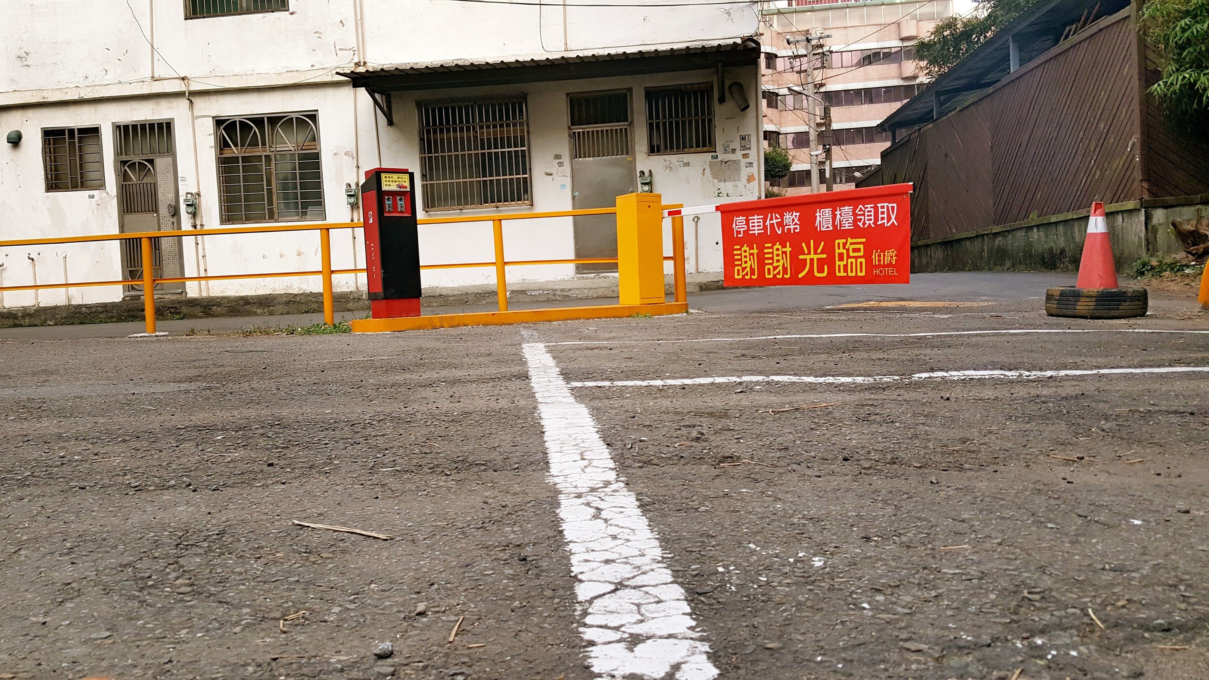Un'area di parcheggio con un cancello giallo, un segnale rosso con il nome dell'hotel e un edificio con grate alle finestre sullo sfondo.