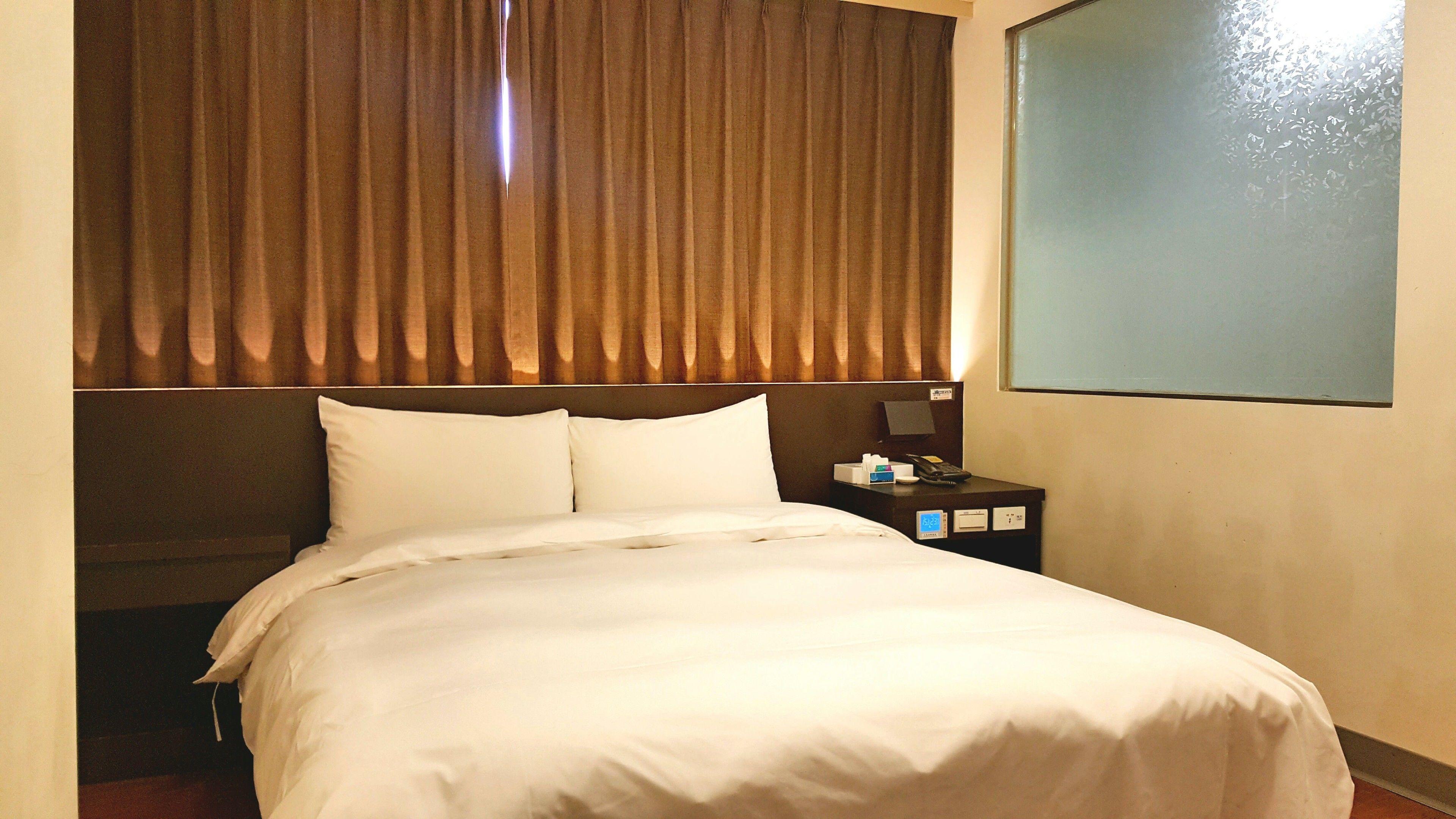 Una habitación de hotel con una cama grande, una mesita de noche con un teléfono y una caja, y una ventana con persianas.