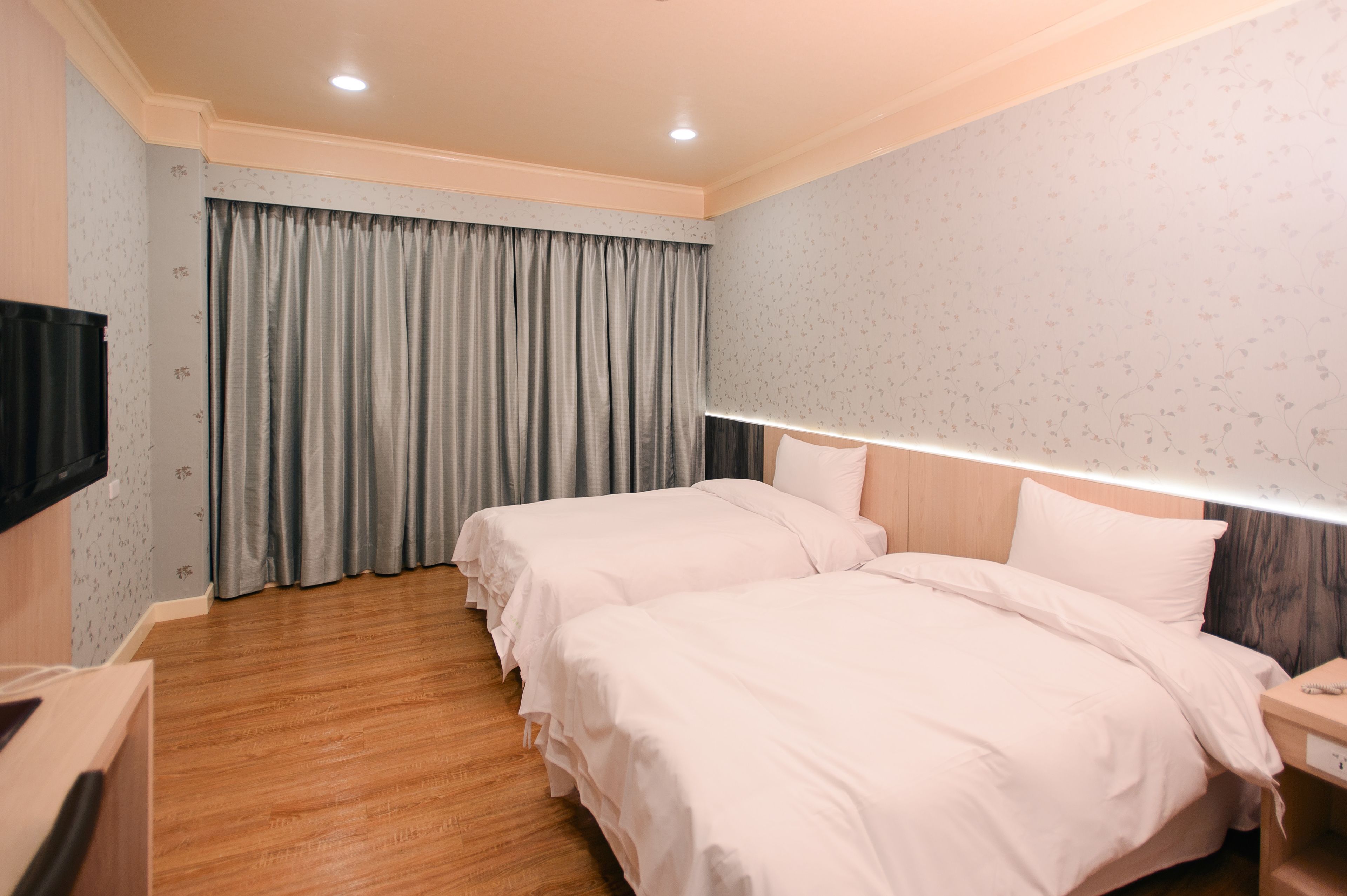 A hotel room with two beds, a TV, a bedside table, and a large window with curtains.