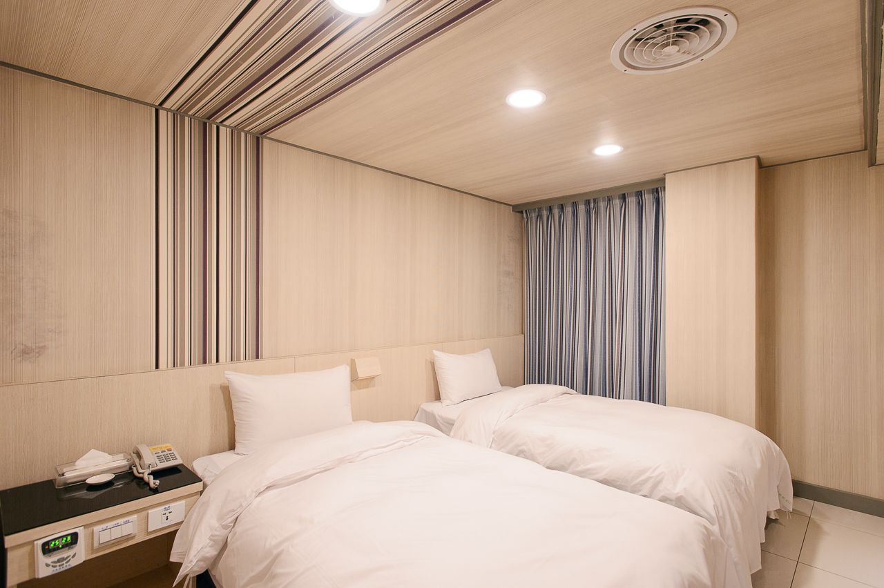 Una habitación de hotel con dos camas, una mesita de noche con teléfono y reloj, y una ventana con cortinas.