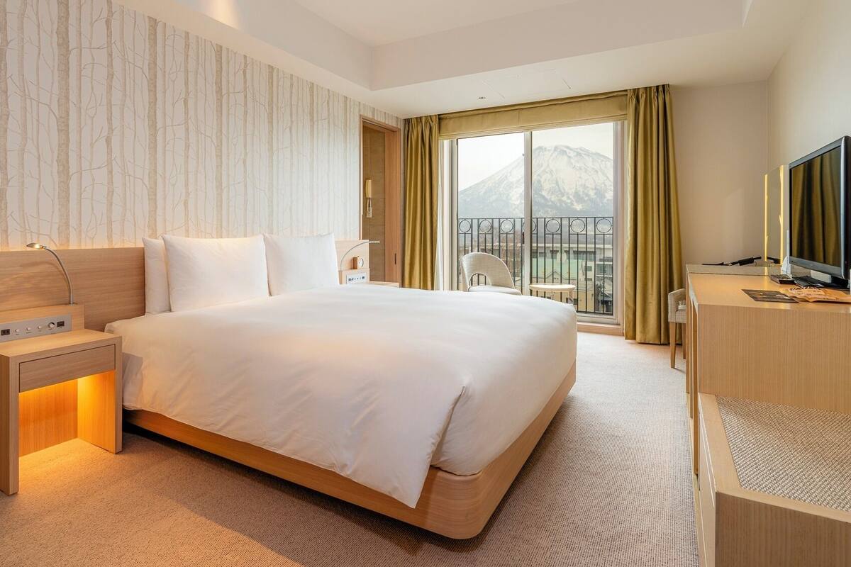A hotel room with a large bed, a desk with a computer, a TV, and a window with a mountain view.