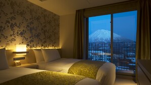 A hotel room with two beds, a nightstand with a lamp, a window with a view of a snow-capped mountain, and a balcony with a railing. - Chalet Ivy Hirafu (Kutchan)
