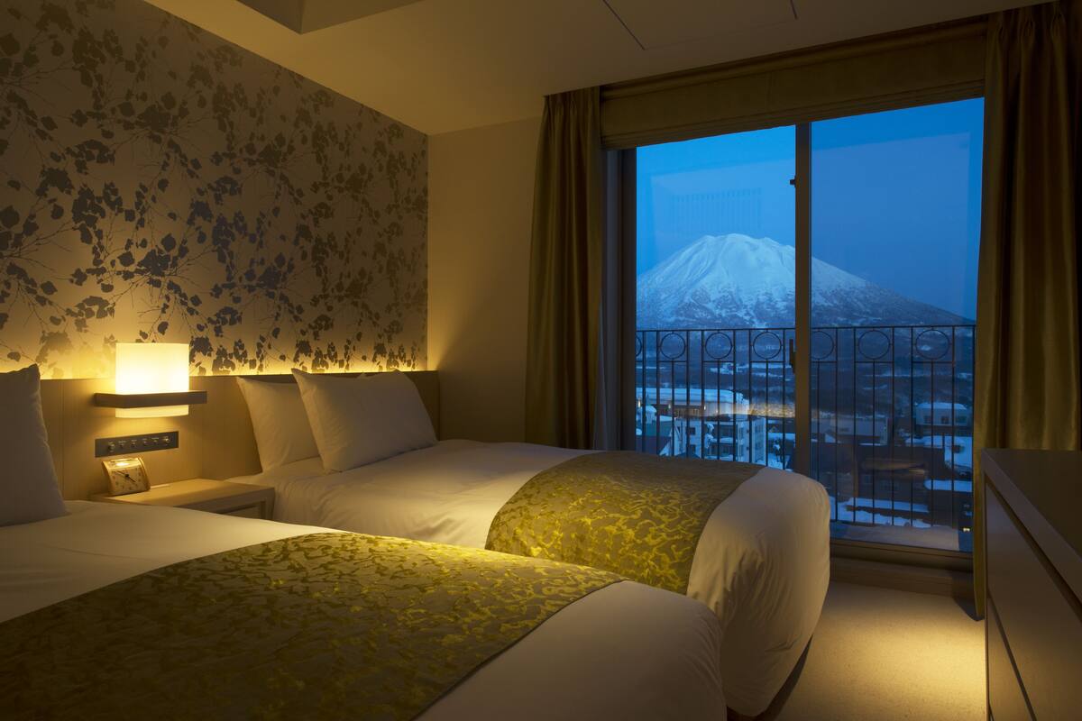 A hotel room with two beds, a nightstand with a lamp, a window with a view of a snow-capped mountain, and a balcony with a railing.