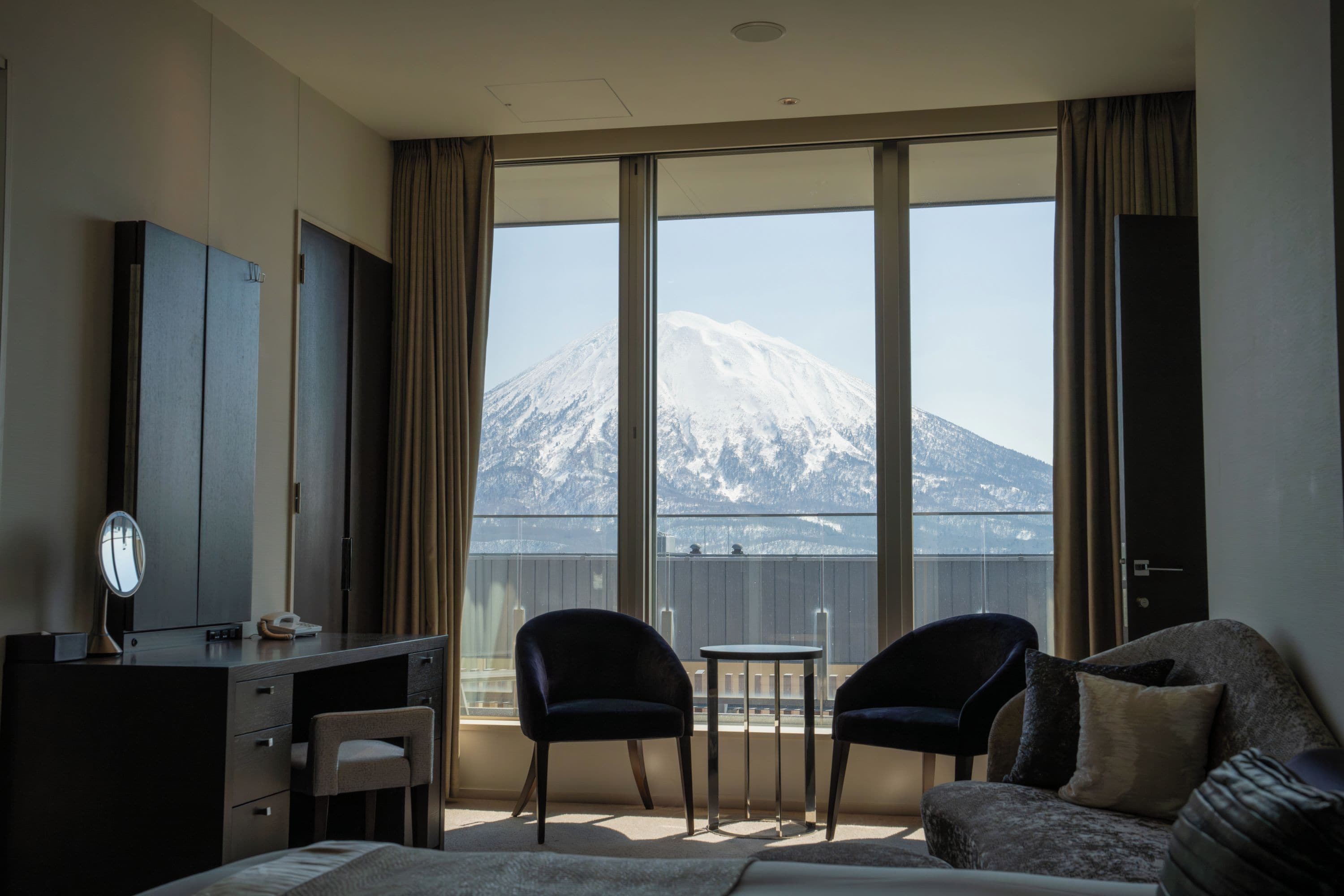 A hotel room with a bed, a desk, a chair, a small table, and a view of a mountain.