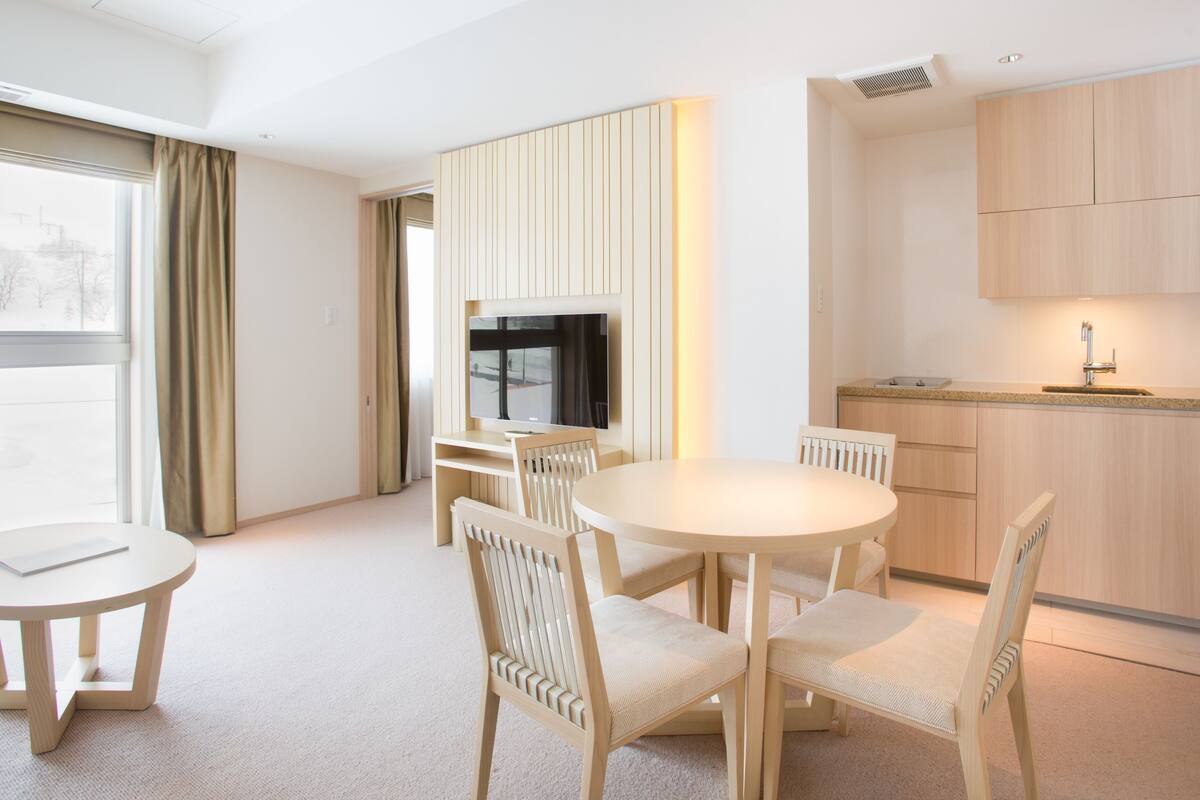 A modern hotel room with a round dining table, four chairs, a TV, a kitchen area with a sink and cabinets, and a large window with curtains.
