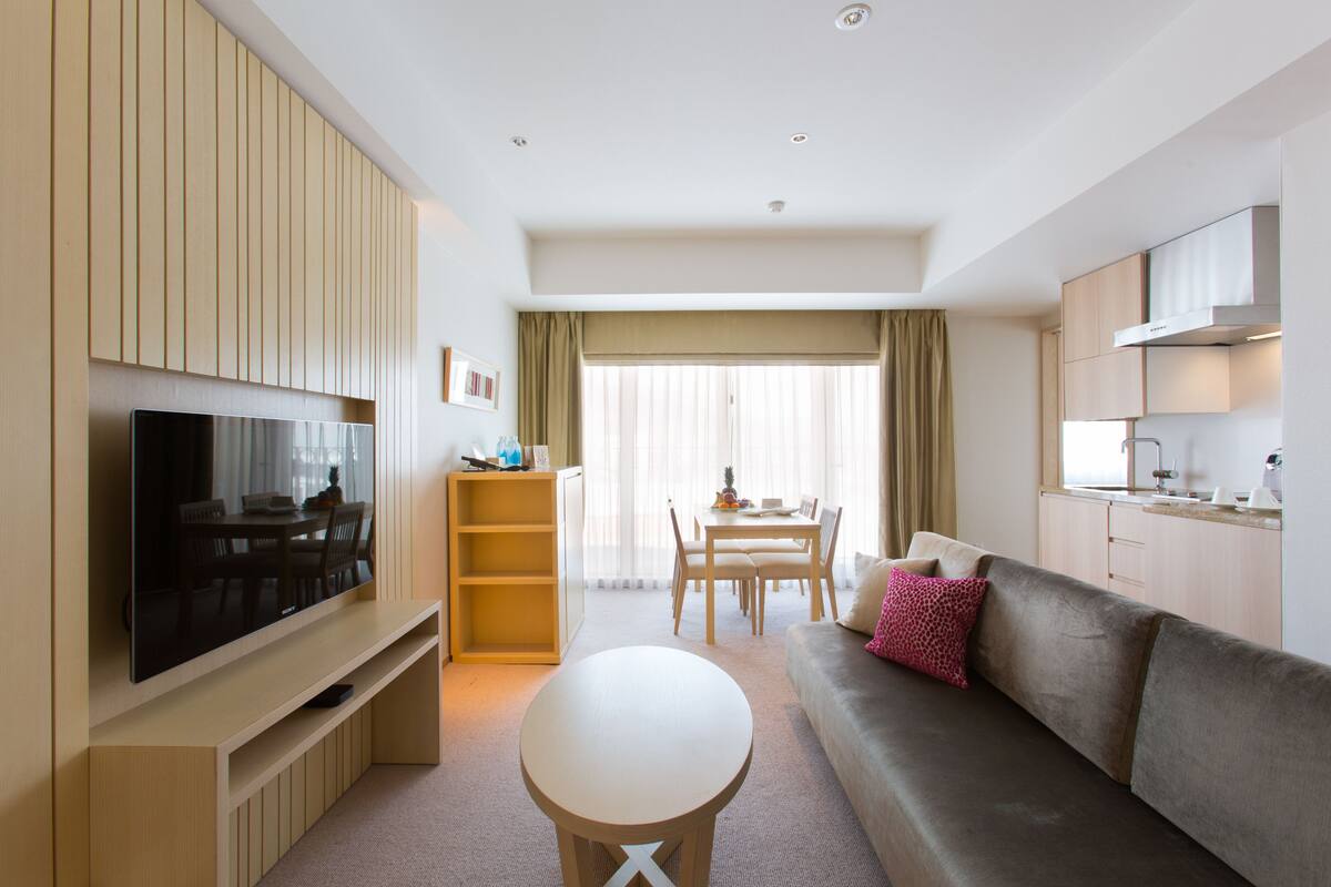 A modern hotel room with a flat-screen TV, a sofa, a dining table, and a kitchen area.