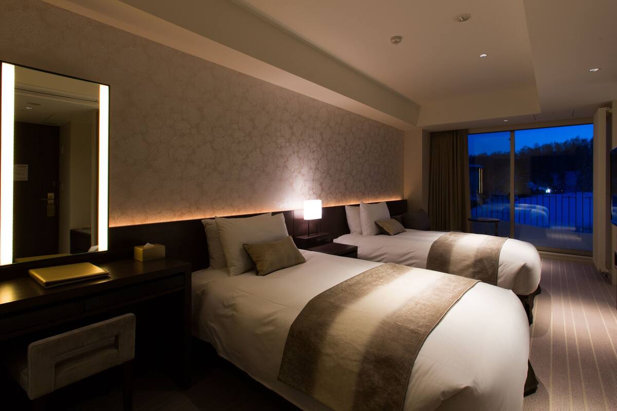 A hotel room with two beds, a large mirror, a desk with a lamp, and a TV.