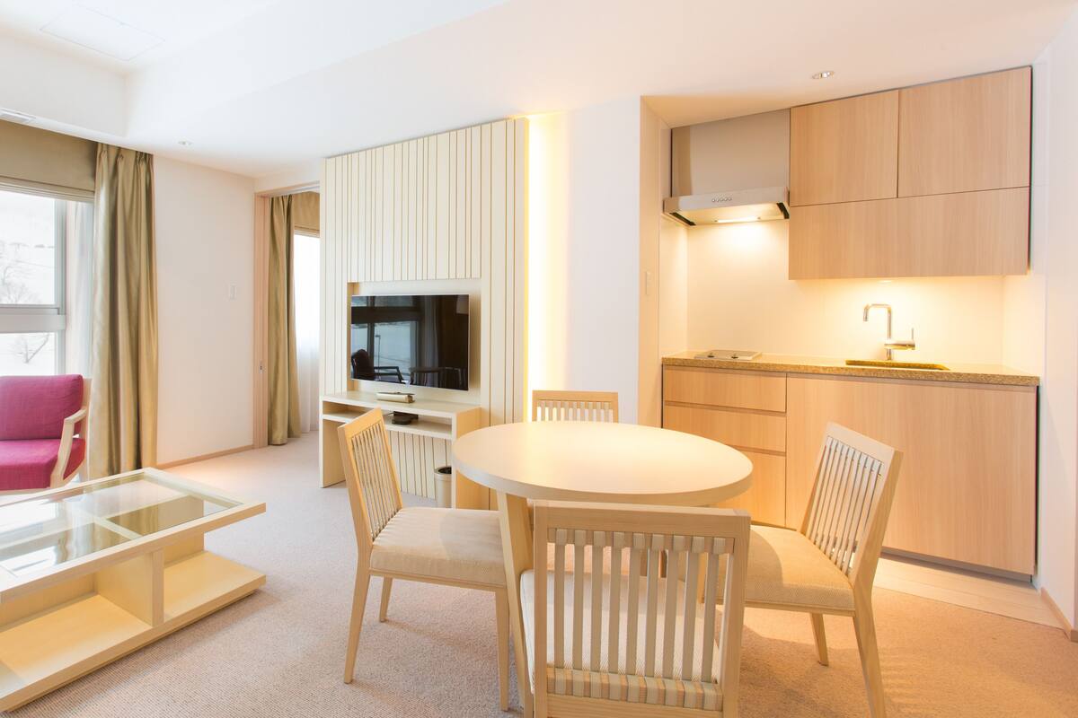 A modern hotel room with a dining area, a TV mounted on the wall, and a kitchen with wooden cabinets.