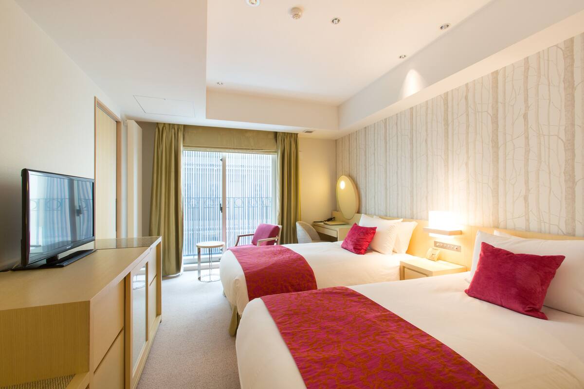 A hotel room with two beds, a desk with a television, a chair, a desk lamp, and a window with curtains.
