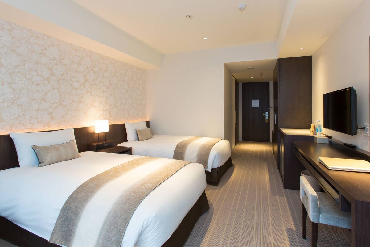 A hotel room with two beds, a desk with a TV, a mirror, and a patterned wallpaper.