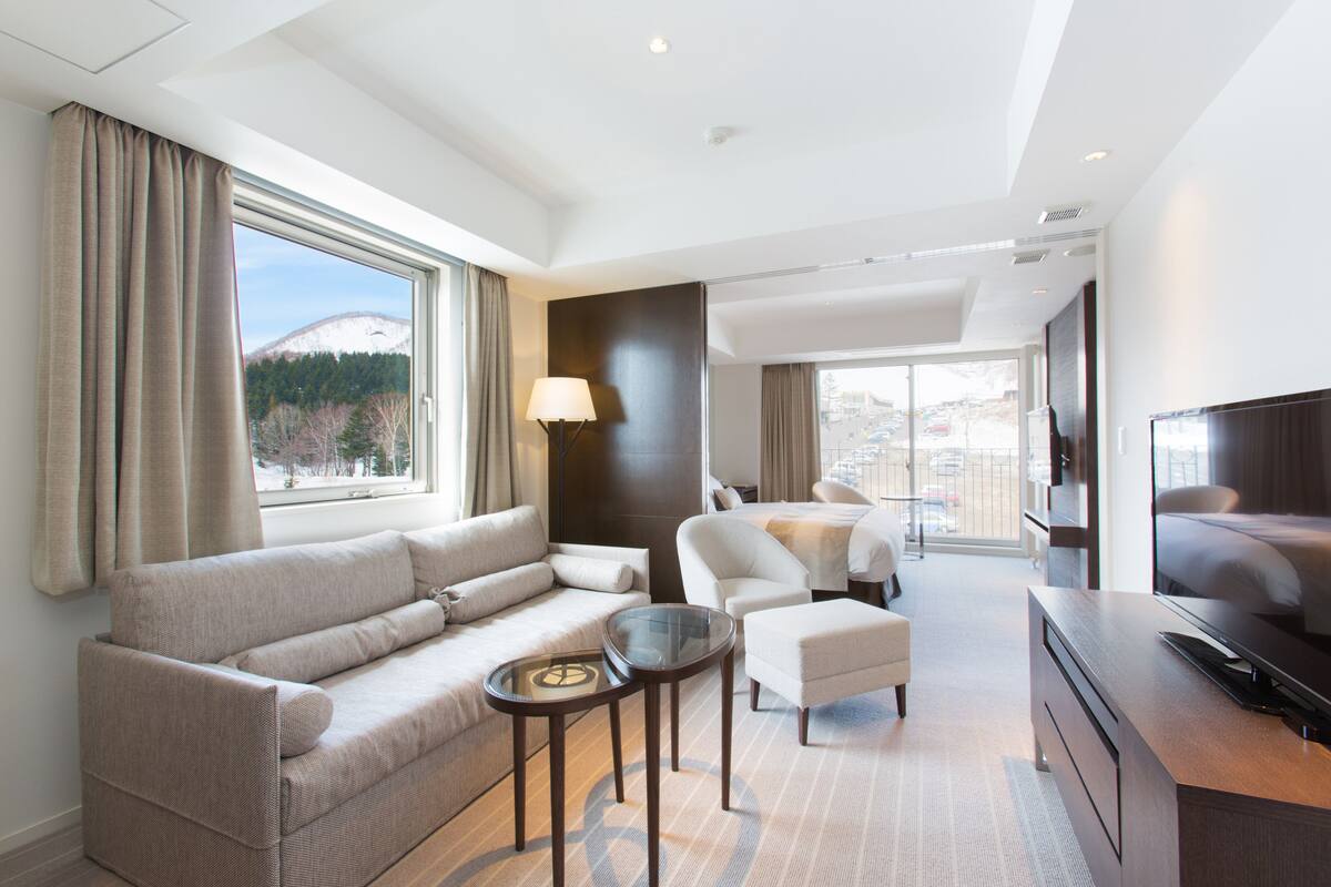 A modern hotel room with a sofa, armchairs, a small table, a TV, and a large window offering a view of a snowy mountain and trees.