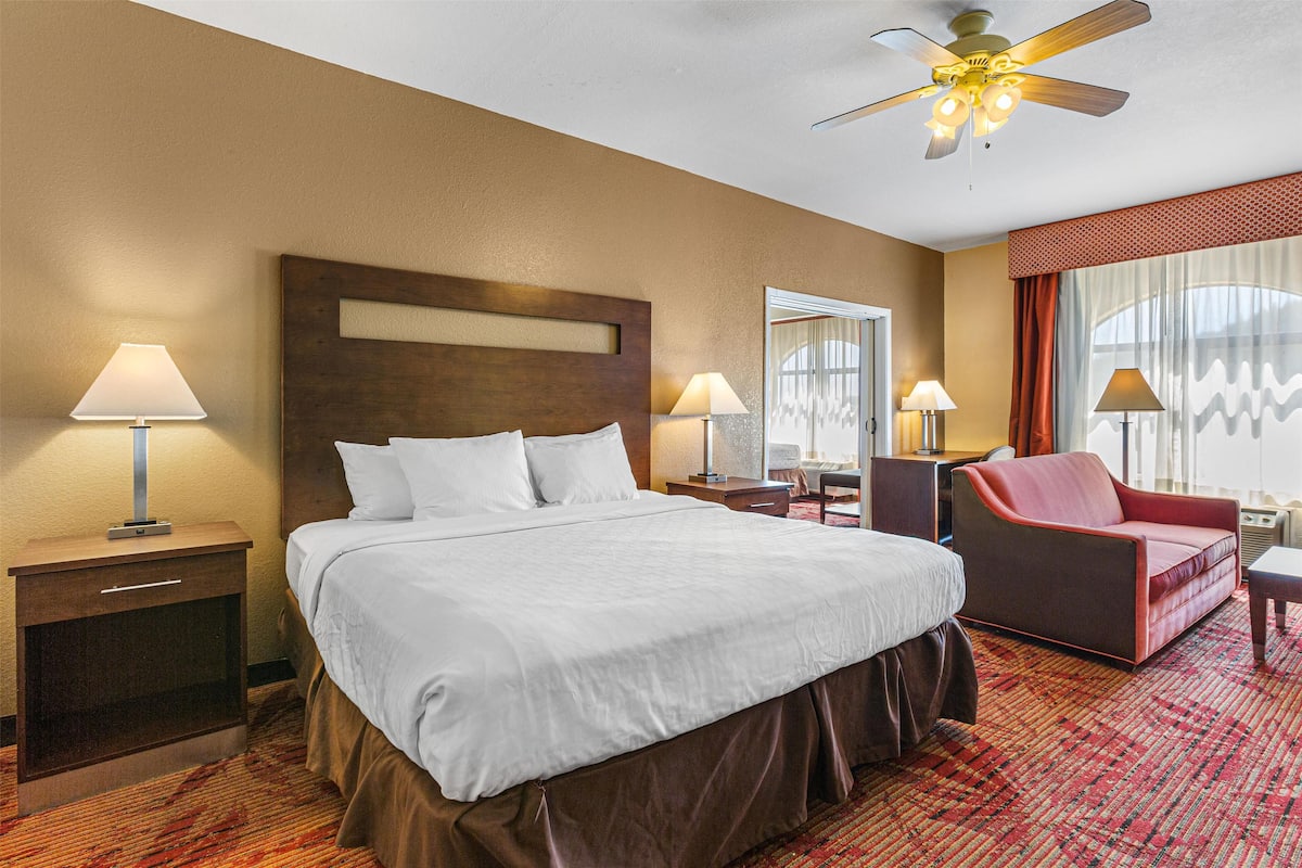A hotel room with a large bed, two bedside tables with lamps, a red armchair, and a ceiling fan.