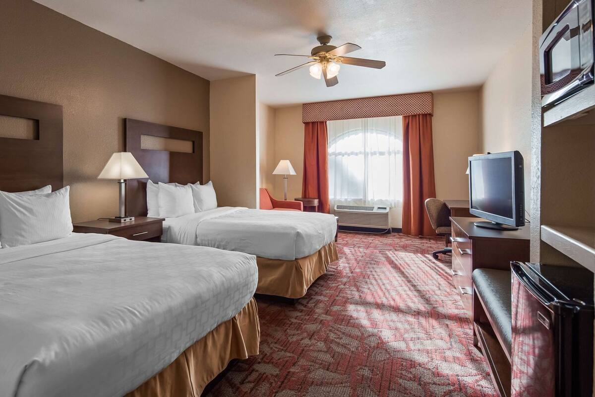 A hotel room with two beds, a television, a desk, a chair, a ceiling fan, and a window with curtains.