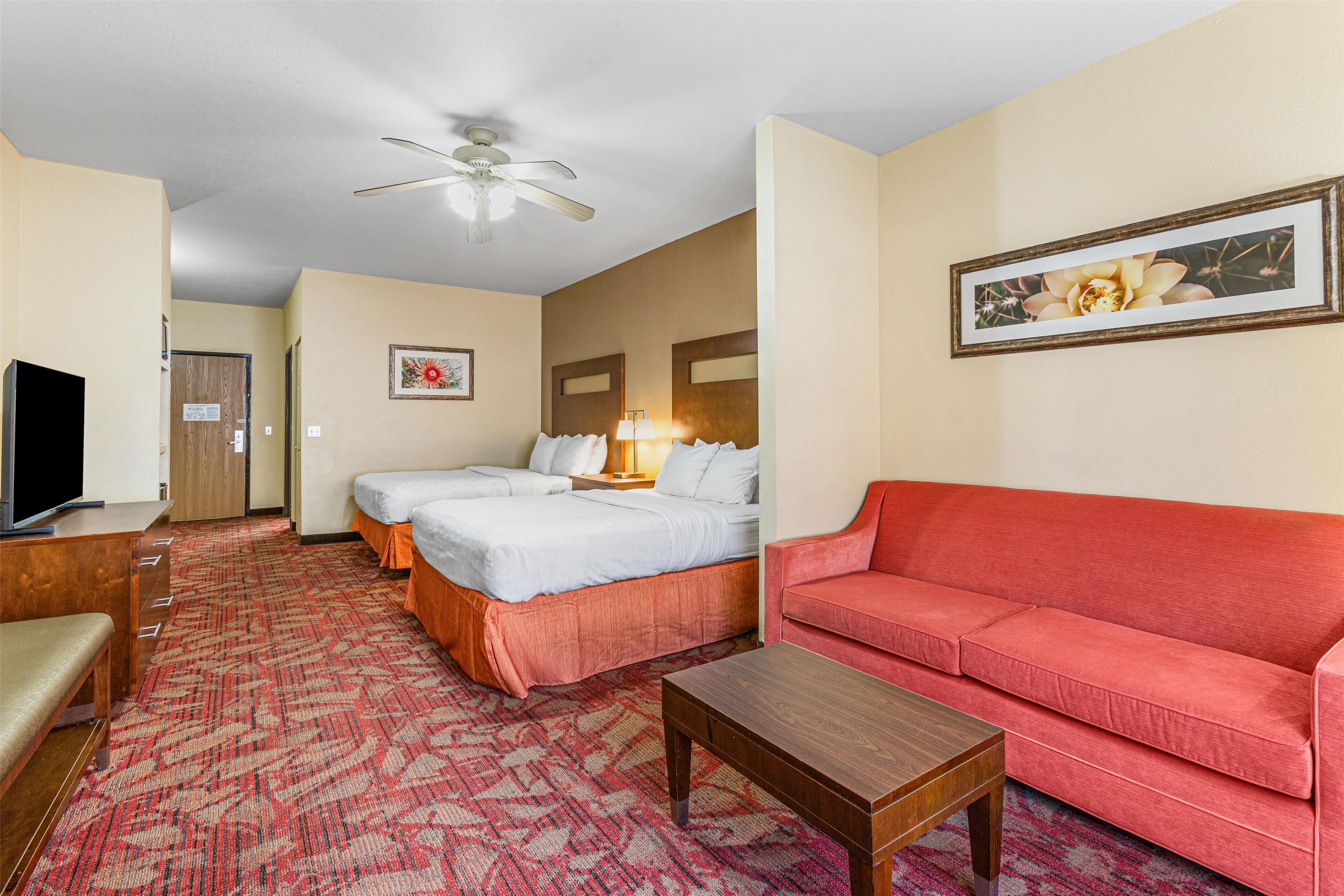 Habitación de hotel con dos camas, un sofá rojo, una mesa de centro de madera, un televisor y un ventilador de techo.