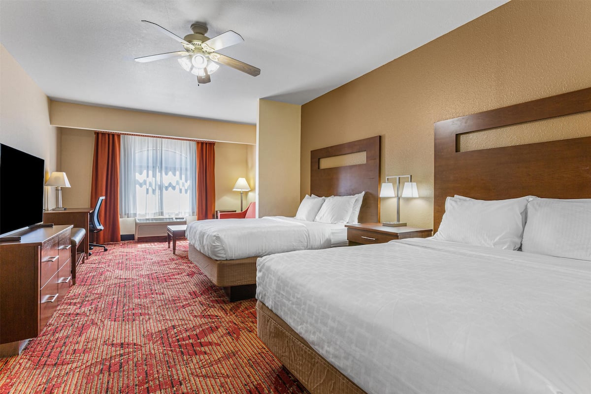A hotel room with two beds, a desk, a chair, a TV, and a ceiling fan.