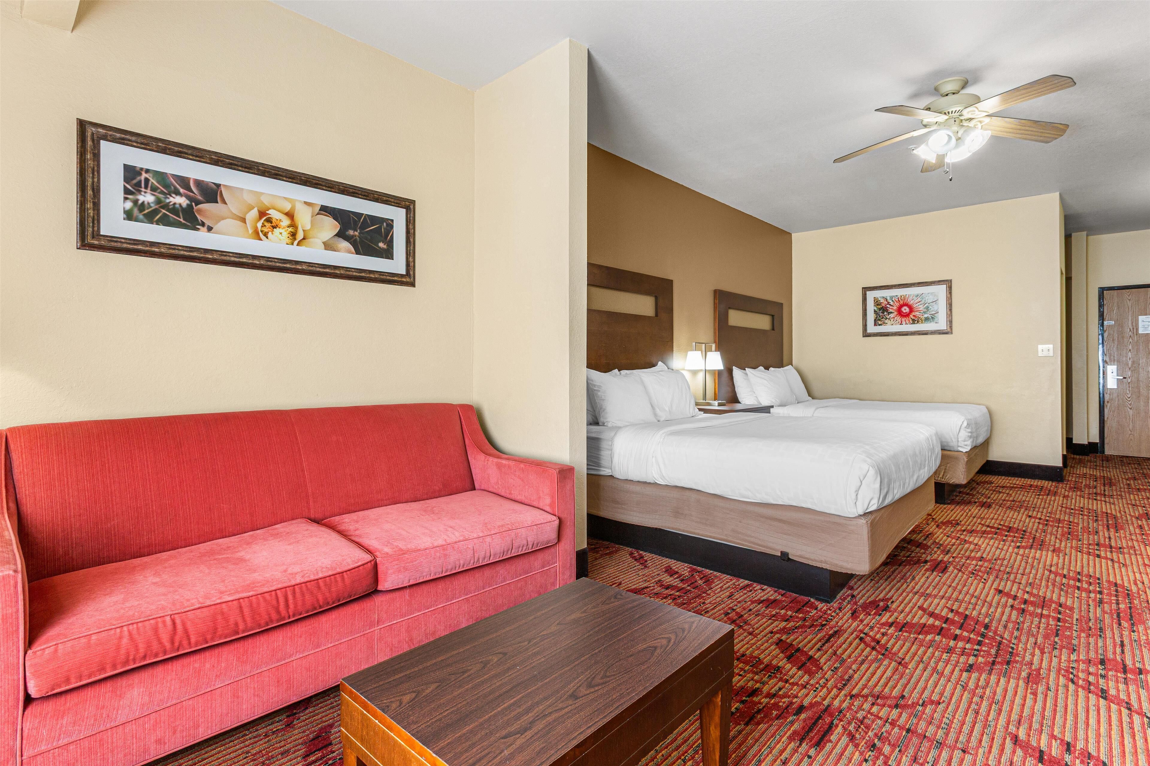 Habitación de hotel con un sofá rojo, una mesa de madera, una cama con ropa blanca y un ventilador de techo.