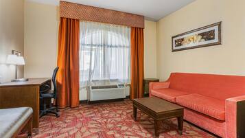 Habitación de hotel con sofá rojo, una mesa de centro de madera, un escritorio con lámpara, aire acondicionado y una alfombra estampada.