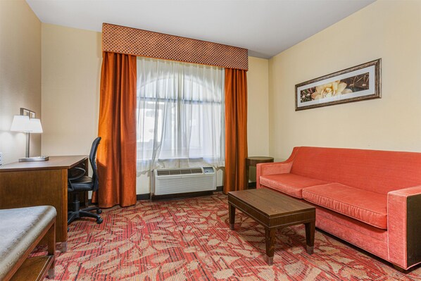 A hotel room with a red sofa, a wooden coffee table, a desk with a lamp, an air conditioner, and a patterned carpet.