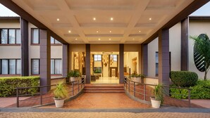 A modern hotel entrance with a brick pathway, potted plants, and a well-lit interior visible through the glass doors.
