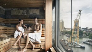 A sauna with wooden benches and a view of a cityscape and a construction crane through a large window. - Haven Hotel Rotterdam, Curio Collection by Hilton (Rotterdam)