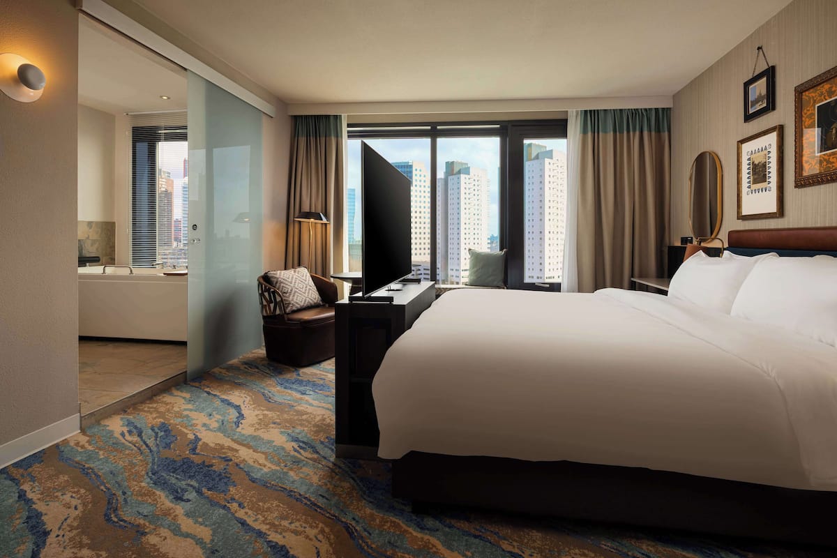 A hotel room with a large bed, a desk with a flat-screen TV, a chair, and a view of tall buildings through the window.