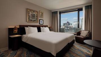 A hotel room with a large bed, a city view, a small table, and a chair.