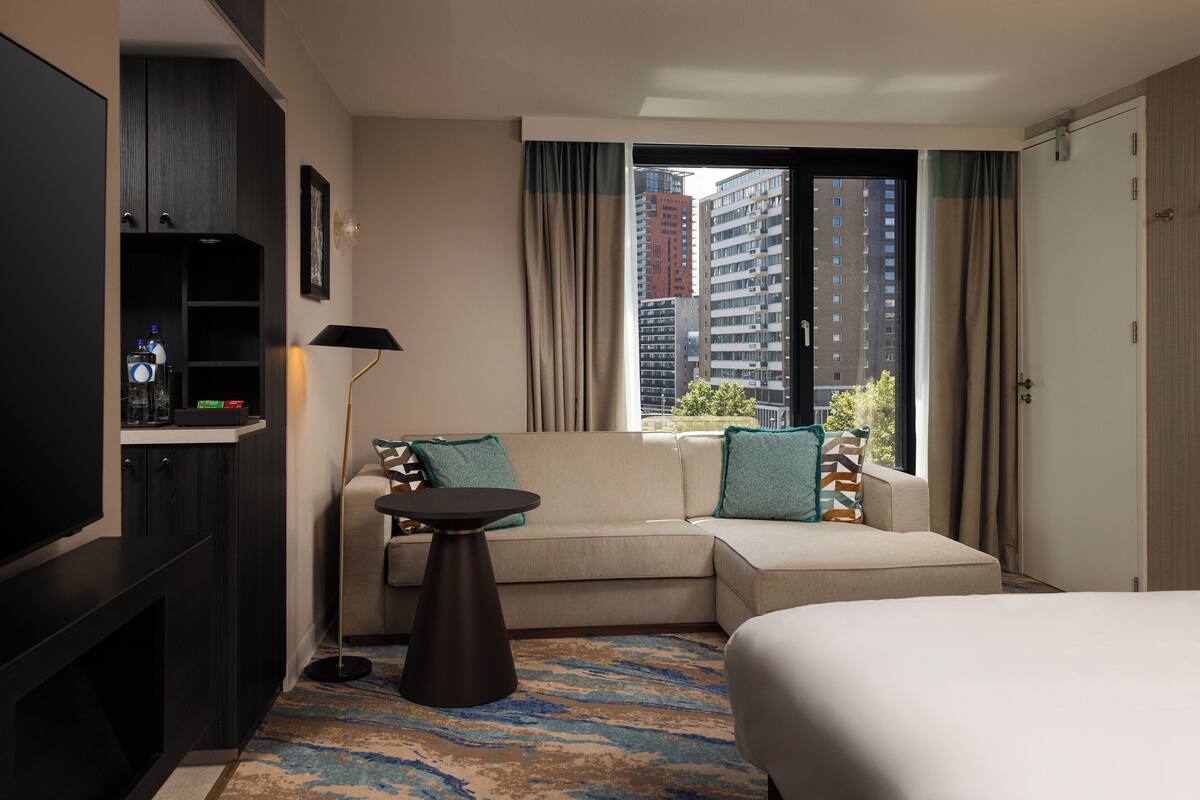 A hotel room with a sofa, a small table, a bed, a TV, and a view of buildings.