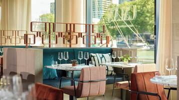 Een restaurantinterieur met een modern design, met een zithoek met tafels en stoelen, een decoratieve metalen scheiding en een uitzicht op de stad door grote ramen.
