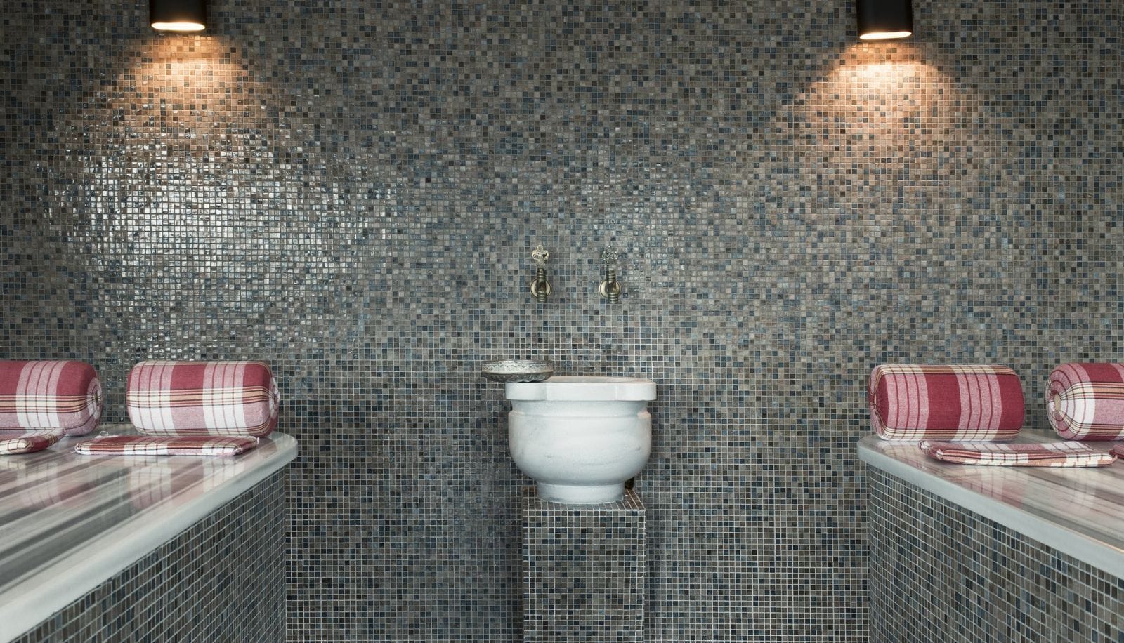 Un baño con paredes de mosaicos, un inodoro blanco y dos toallas enrolladas sobre un mostrador.