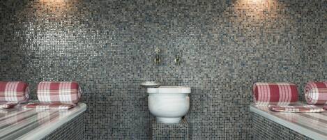 Un baño con paredes de mosaicos, un inodoro blanco y dos toallas enrolladas sobre un mostrador.