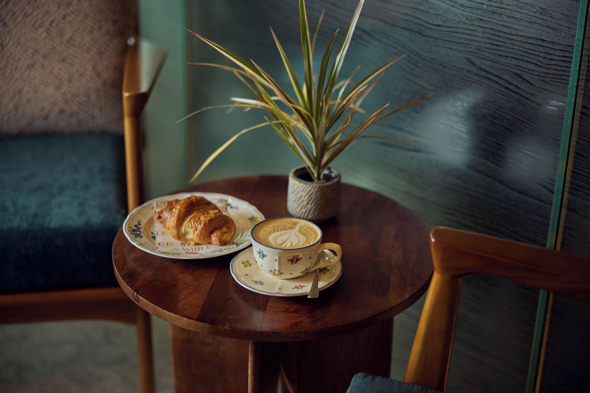 A round wooden table with a cup of coffee, a plate with a croissant, and a potted plant.