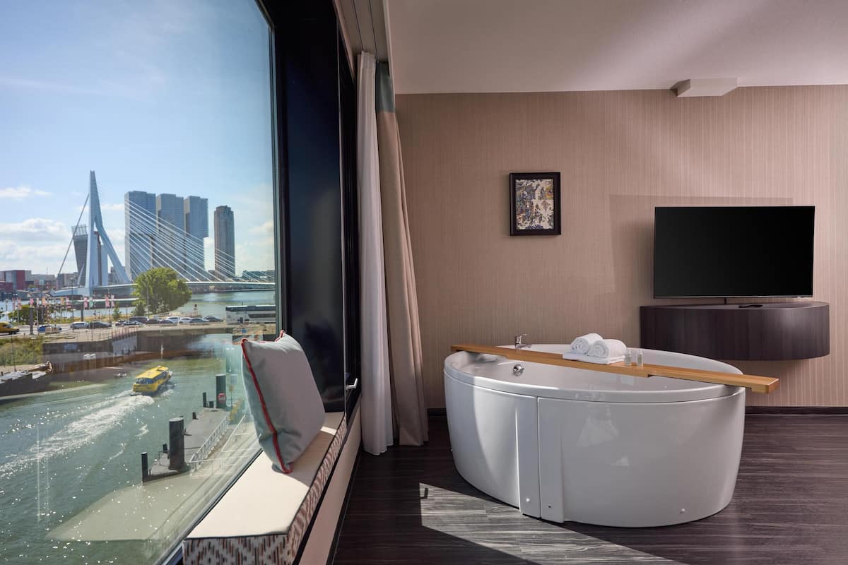 A modern hotel room with a large window offering a city view, a seating area with a view, a flat-screen TV, and a small table with towels.