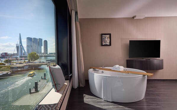 A modern hotel room with a large window offering a city view, a seating area with a view, a flat-screen TV, and a small table with towels.