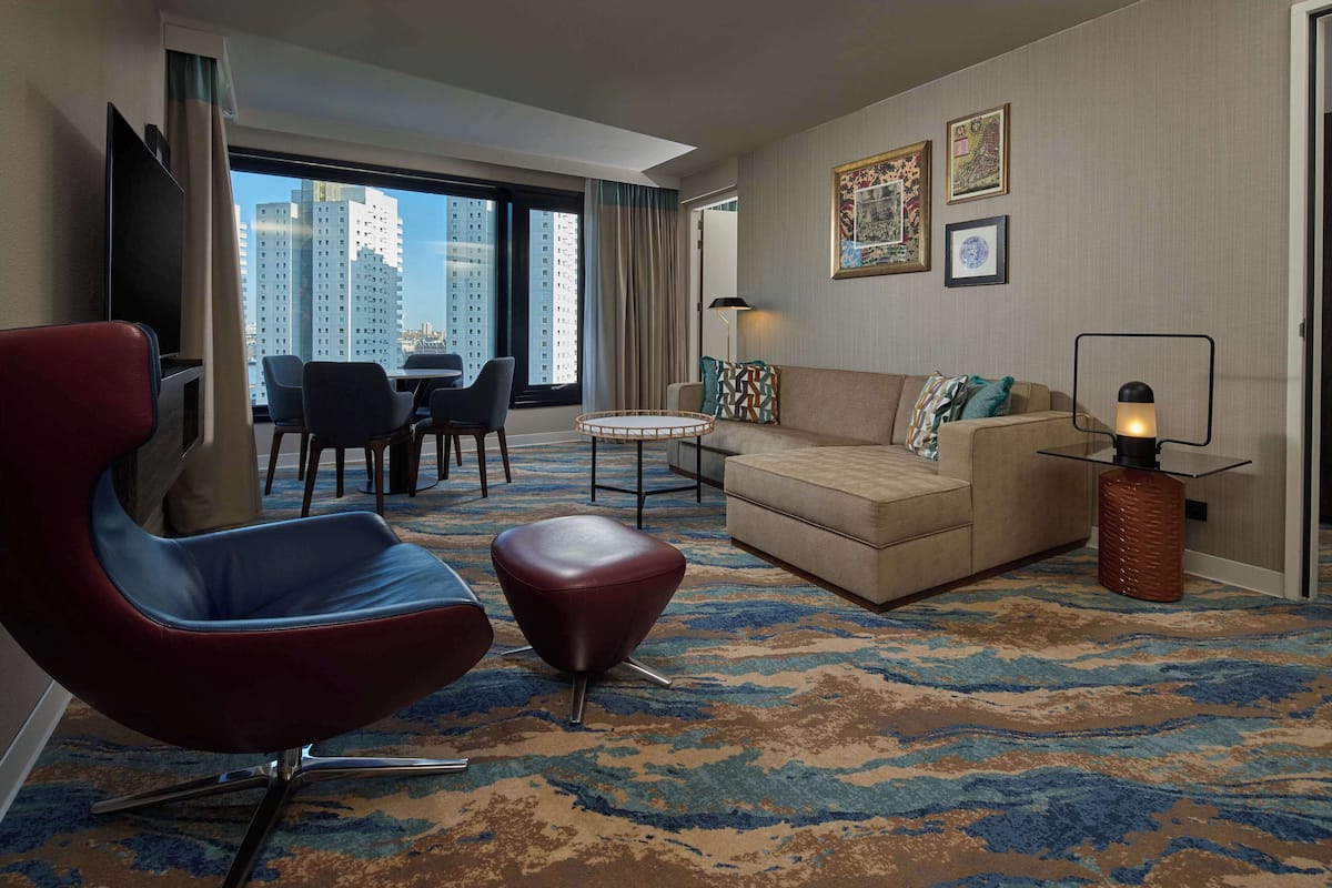 A hotel room with a modern living area, including a red leather chair, a beige sofa, a small coffee table, and a patterned carpet.