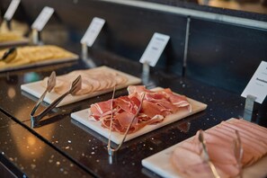 A buffet with various food items, including sliced meats and cheeses, displayed on white plates with metal tongs.