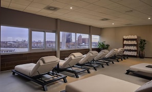 A room with multiple reclining chairs, a large window with a city view, and a shelf with rolled towels.