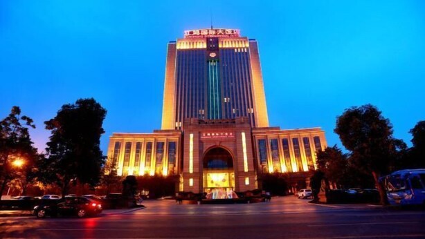 Un edificio grande e iluminado con una entrada central y columnas verticales, rodeado de árboles y coches estacionados.