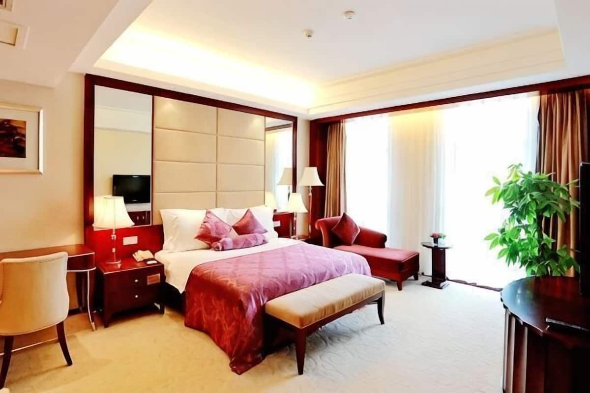 Une chambre d’hôtel avec un grand lit, des tables de chevet, un bureau, une chaise, une plante et une fenêtre avec des rideaux.