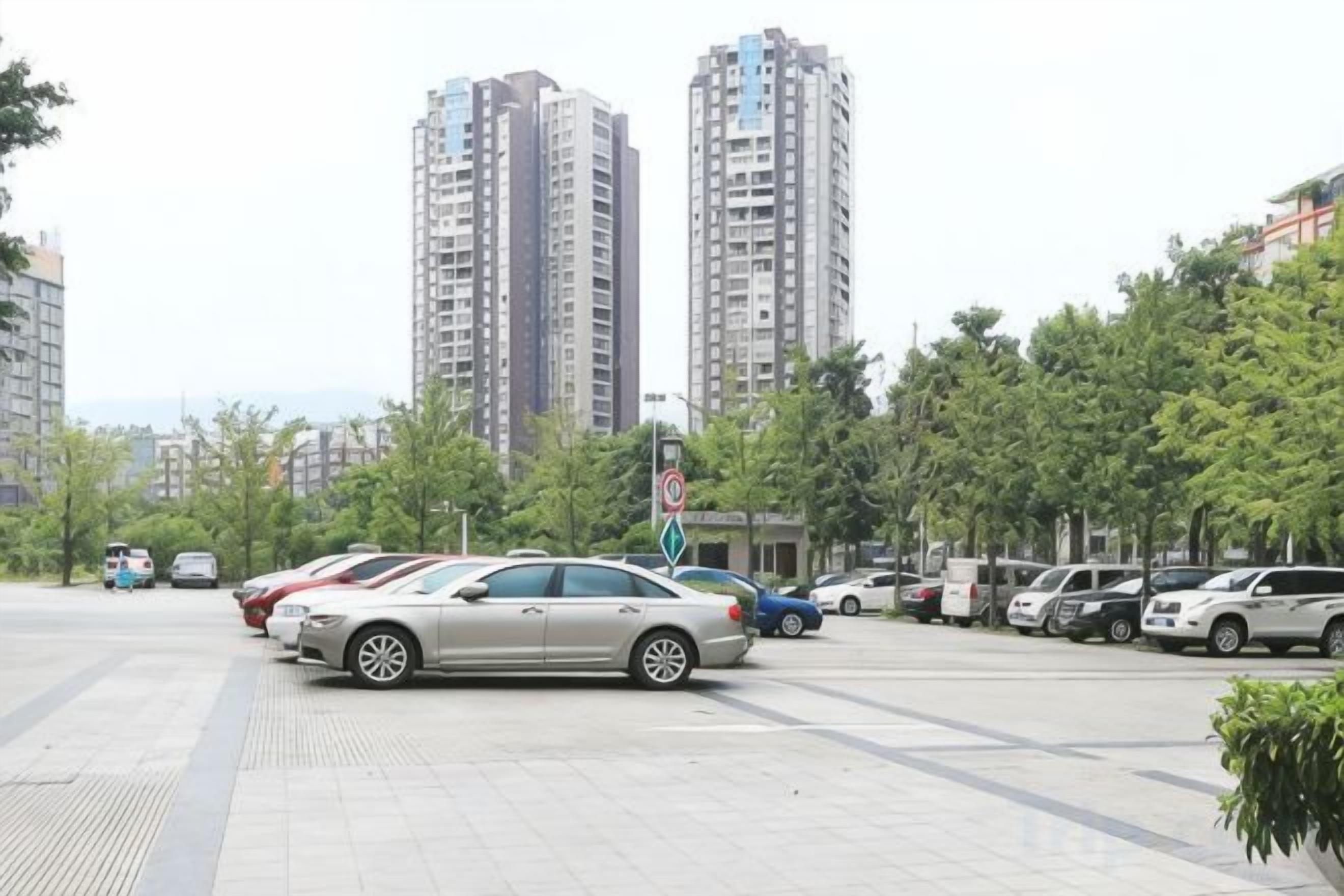 A parking area with multiple cars, tall residential buildings, and trees in the background.