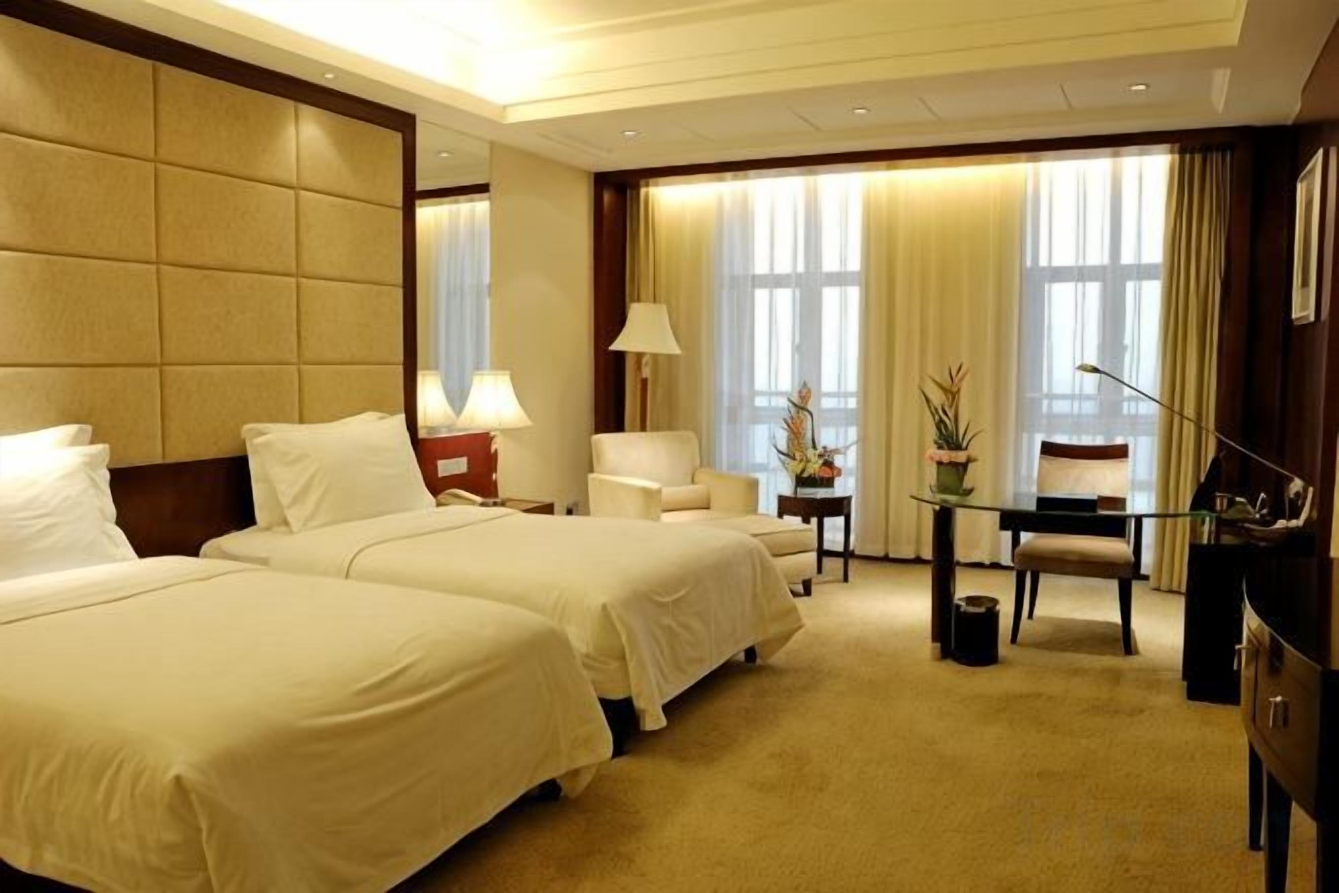 A hotel room with two beds, a desk, a chair, a lamp, and a window with curtains.